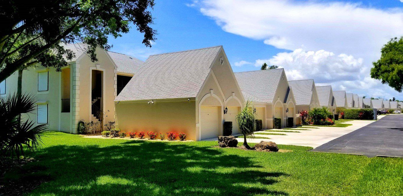 Row of tan houses with white roof, green lawn, and blue sky with clouds.