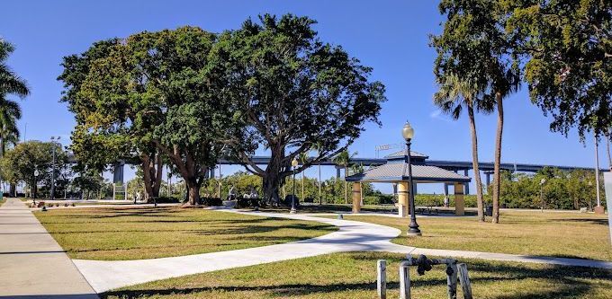 Park with large trees, a paved path, gazebo, and a bridge in the background under a blue sky.