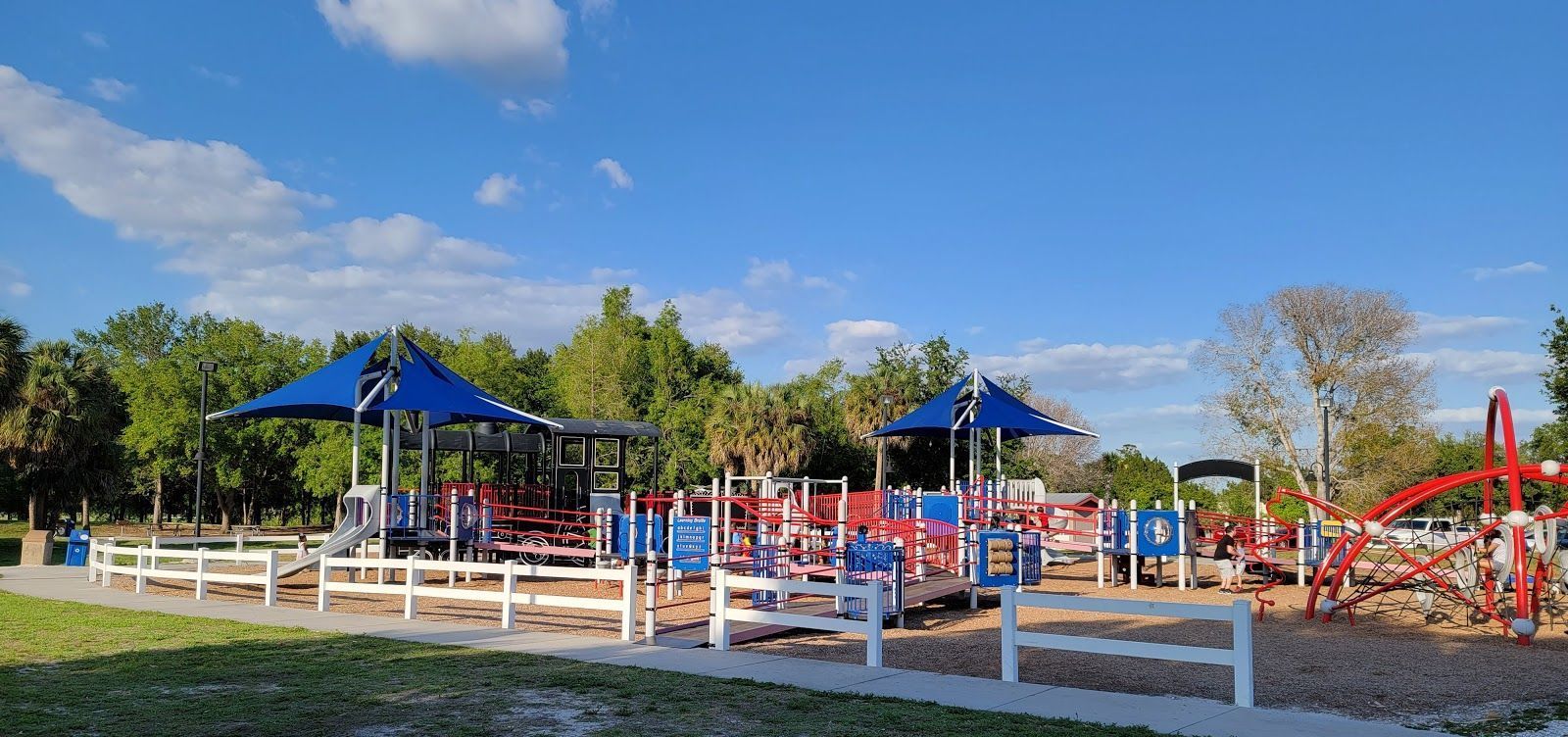 Playground with red, white, and blue equipment, blue sky, trees in the background, and white fence.