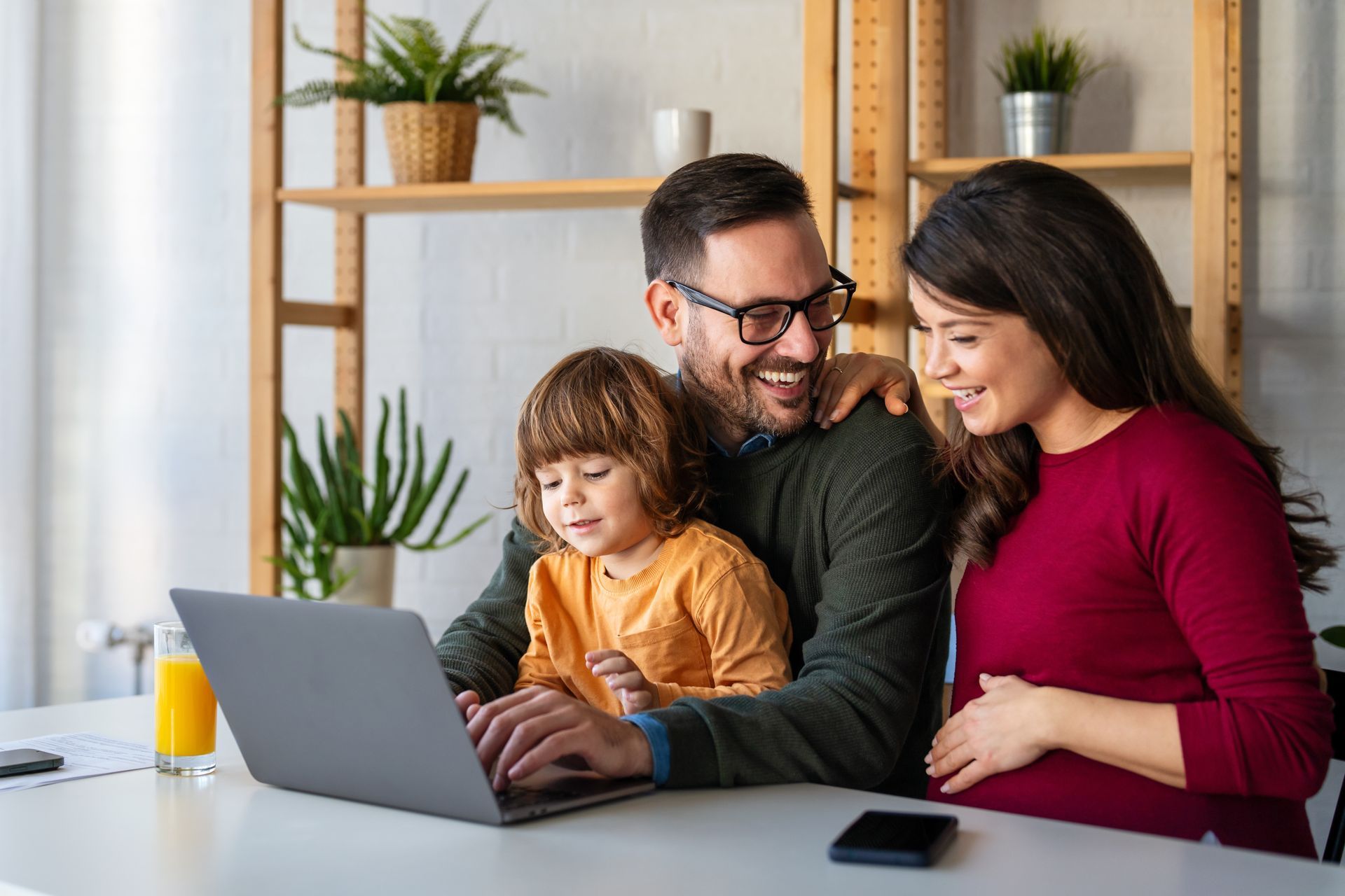Family gathered around a laptop at a table in a bright home office, smiling and chatting
