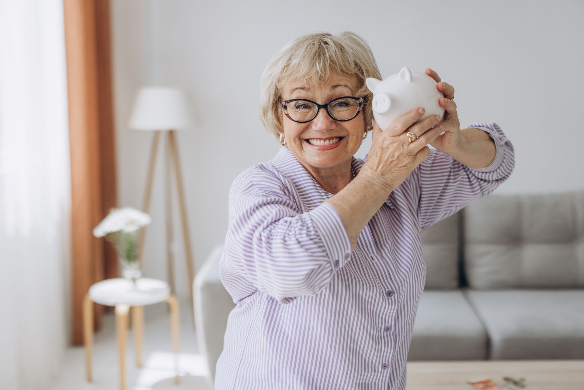 Smiling person with glasses holds a white piggy bank in a bright living room.