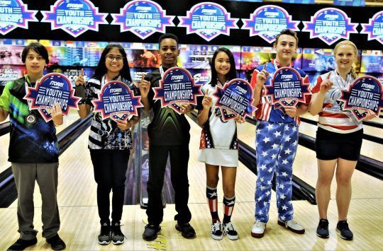 Youth bowling champions hold awards at a bowling alley, celebrating their win.