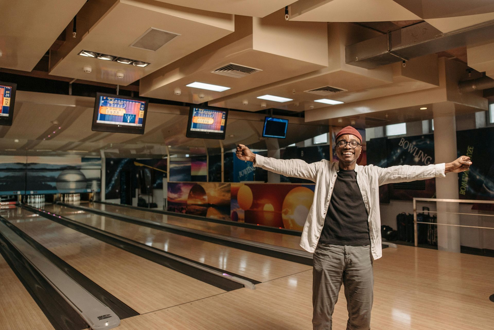 Person with arms outstretched in a bowling alley, smiling. Bowling lanes and scoring screens are visible.