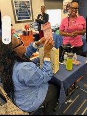 Person seated at a table holds up a pink package while others watch in a community room.