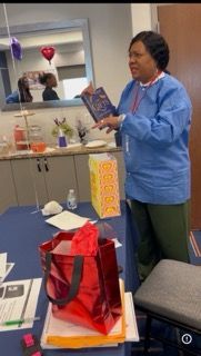 Person in blue shirt holds a book at a table with gift bags in a small office setting.