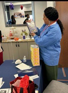 Person in a blue shirt holds a gift bag in a room with balloons, tables, and wrapped presents.