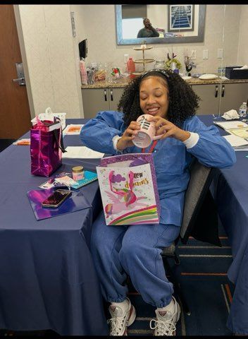 Person smiling on a salon chair, opening a gift bag with presents on a table beside them.