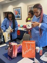 Two people in blue aprons pack gift bags at a table with cups and colorful paper bags.