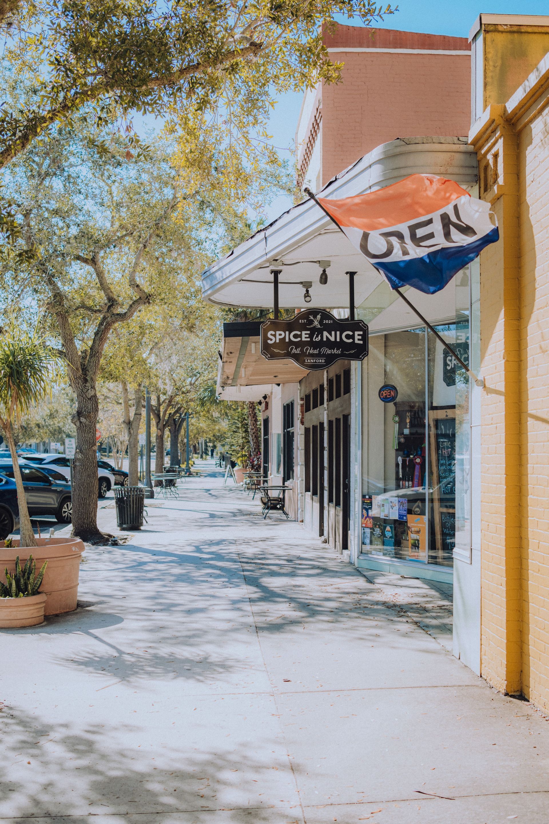 Sunny sidewalk lined with shops and trees, with a white awning and a UTEP sign in the foreground.