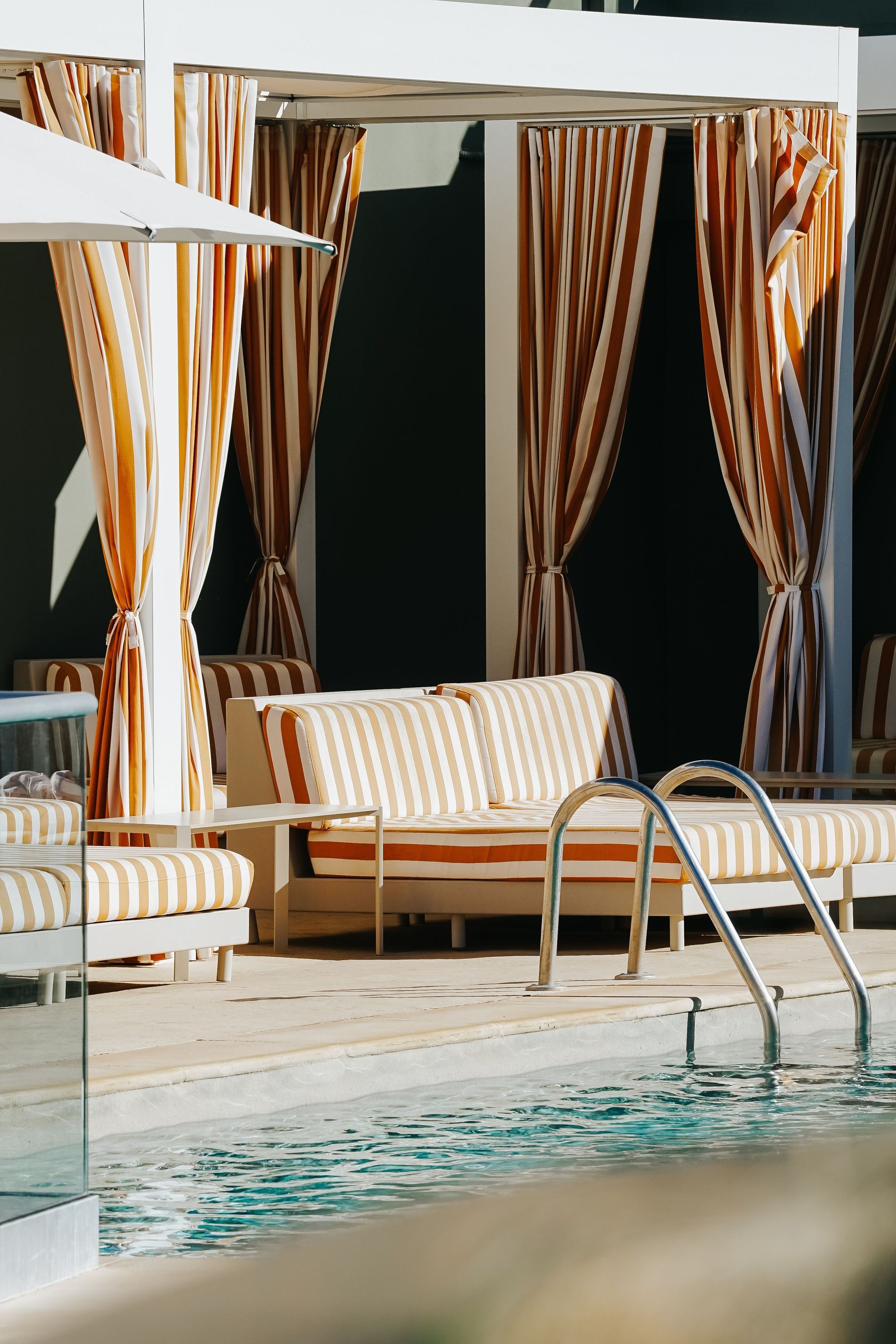 Poolside lounge area with striped curtains, cushioned benches, and a metal ladder by the water