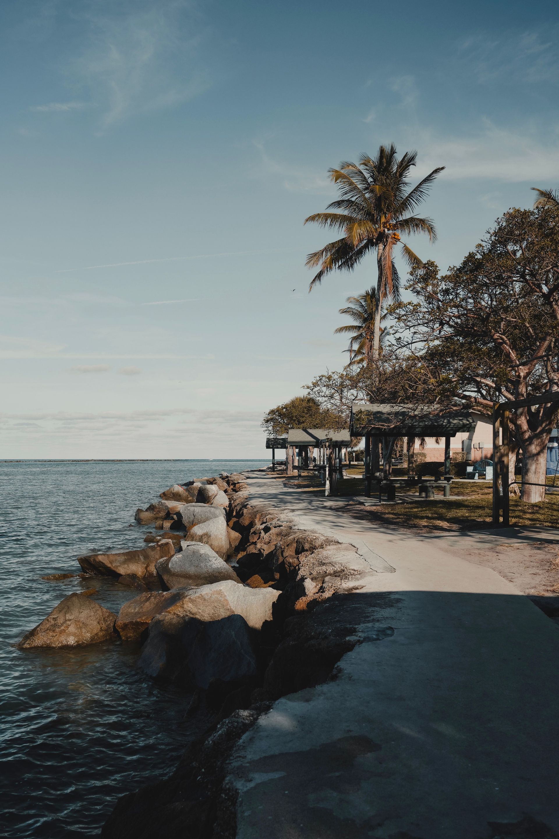 Rocky seaside path beside calm water, palm trees, and a small thatched hut at sunset