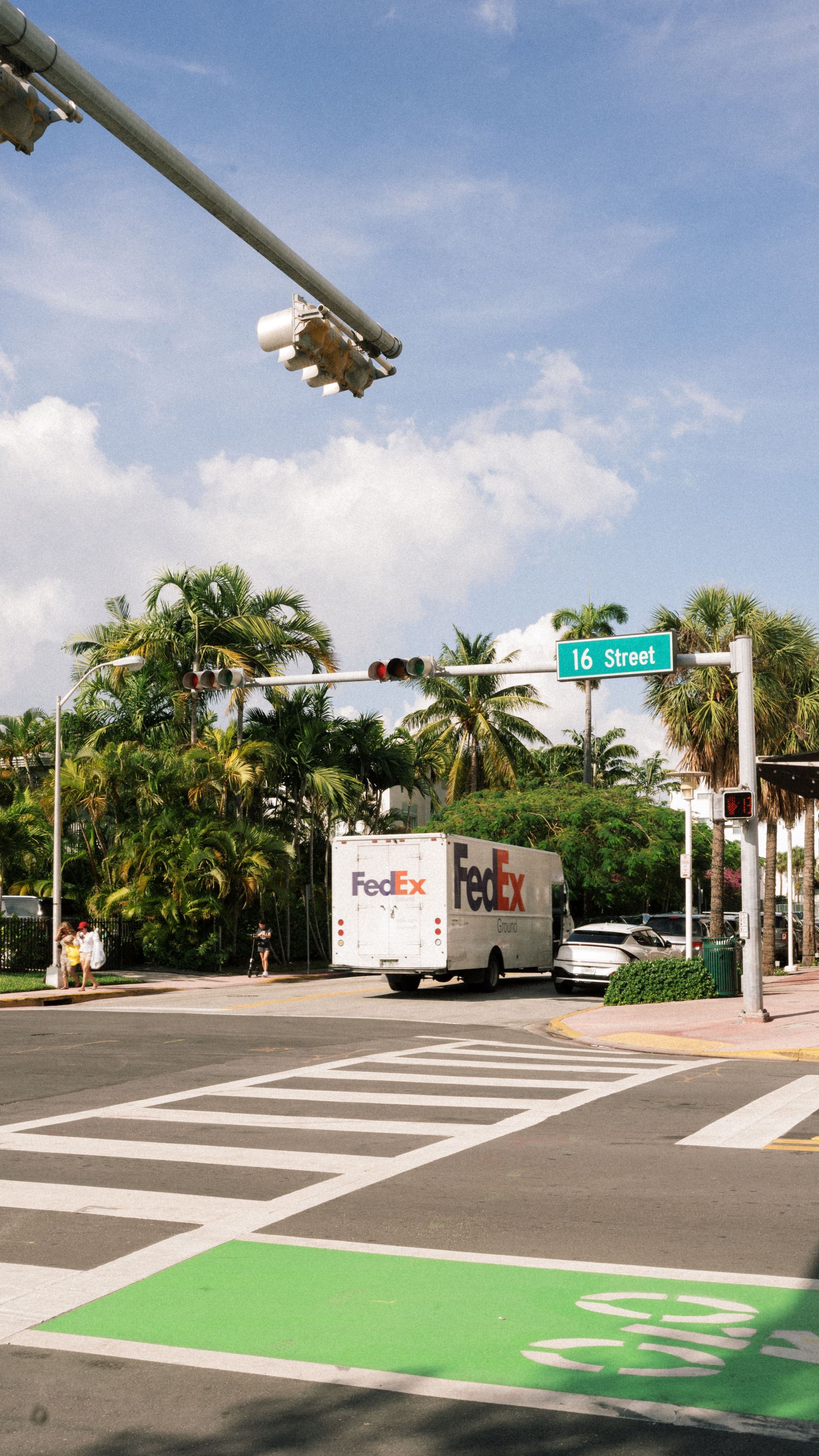 FedEx truck stopped at a crosswalk under a traffic light on a sunny street corner.