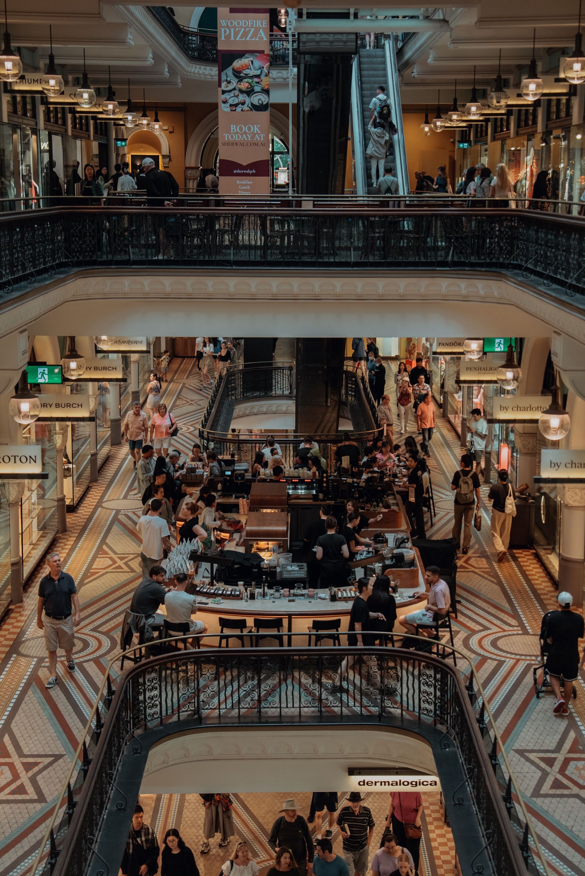Multi-level shopping arcade with ornate railings, tiled floors, and people seated in a central café area.
