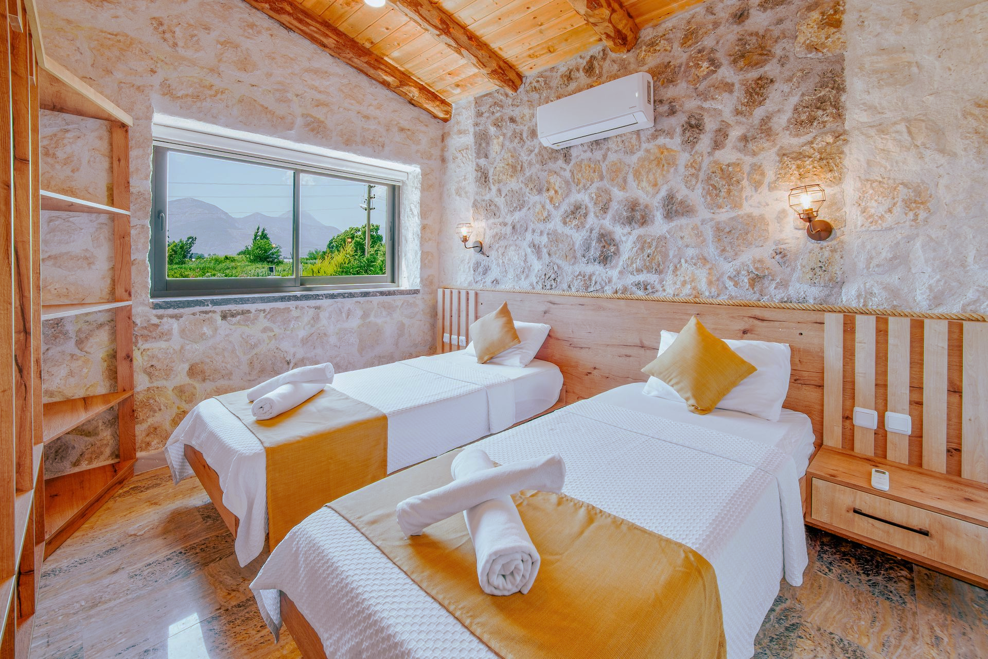 Stone-walled bedroom with two twin beds, yellow accents, and a large window overlooking greenery