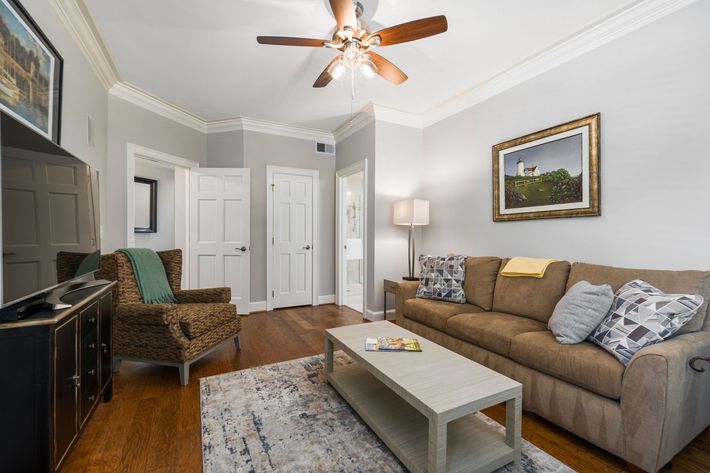 Bright living room with tan sofa, patterned chair, coffee table, lamp, TV, and ceiling fan on gray walls