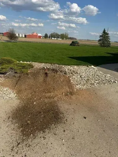 Small mound of dirt and gravel on a concrete surface beside a grassy field under a cloudy sky