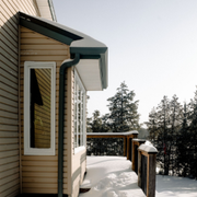 Snow-covered wooden deck beside beige house with green awning and forest beyond