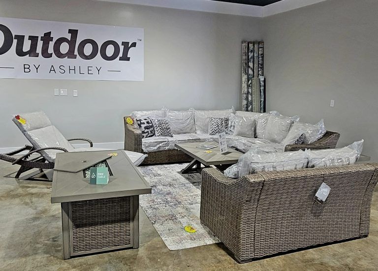 Outdoor furniture showroom with wicker sofa, chairs, coffee table, and rug in a gray display area