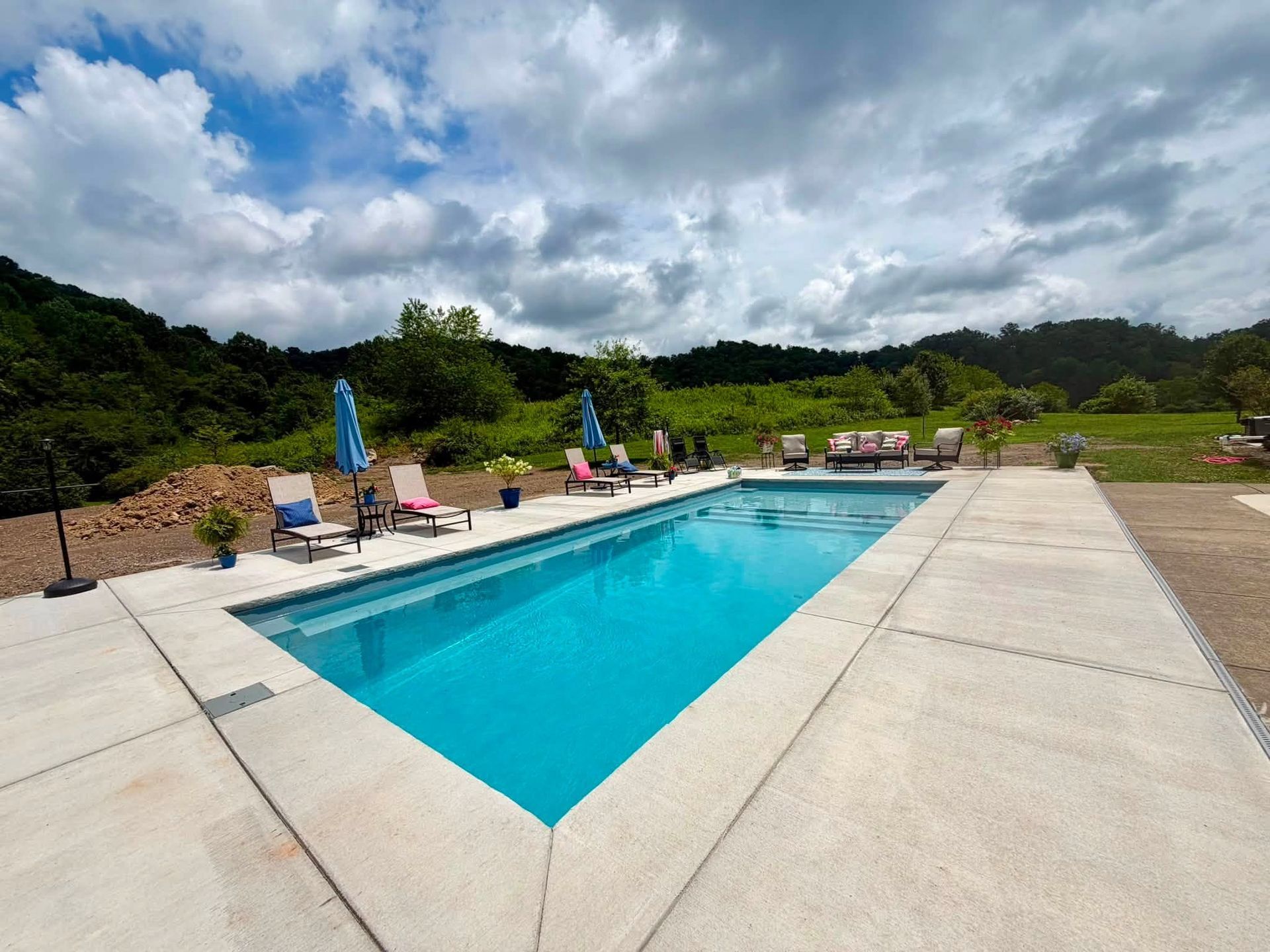 Outdoor swimming pool with turquoise water beside a concrete deck under a cloudy sky, surrounded by green hills.