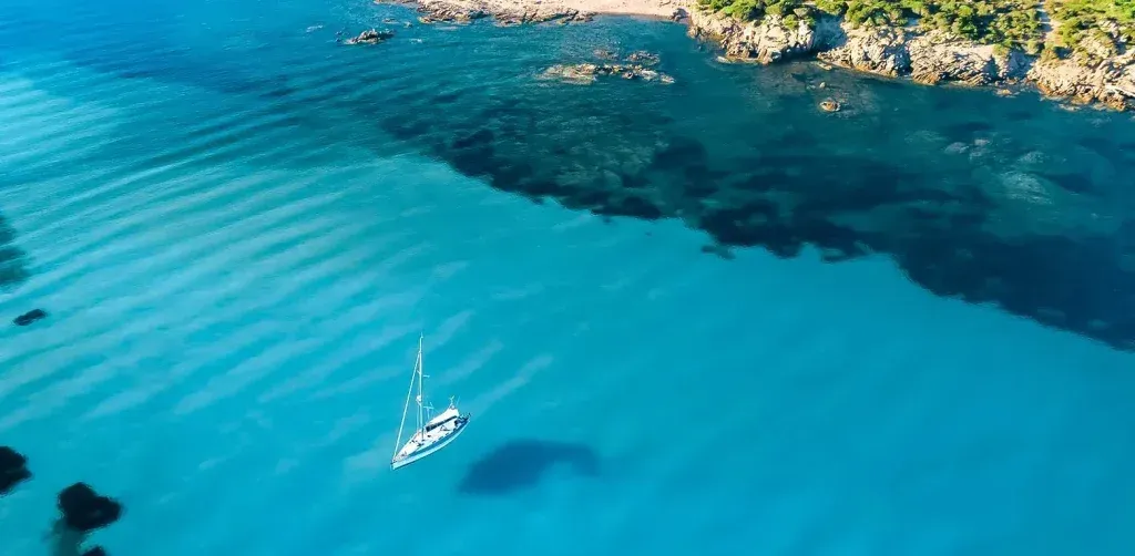 Aerial view of a white boat on bright blue ocean near a rocky coastline.