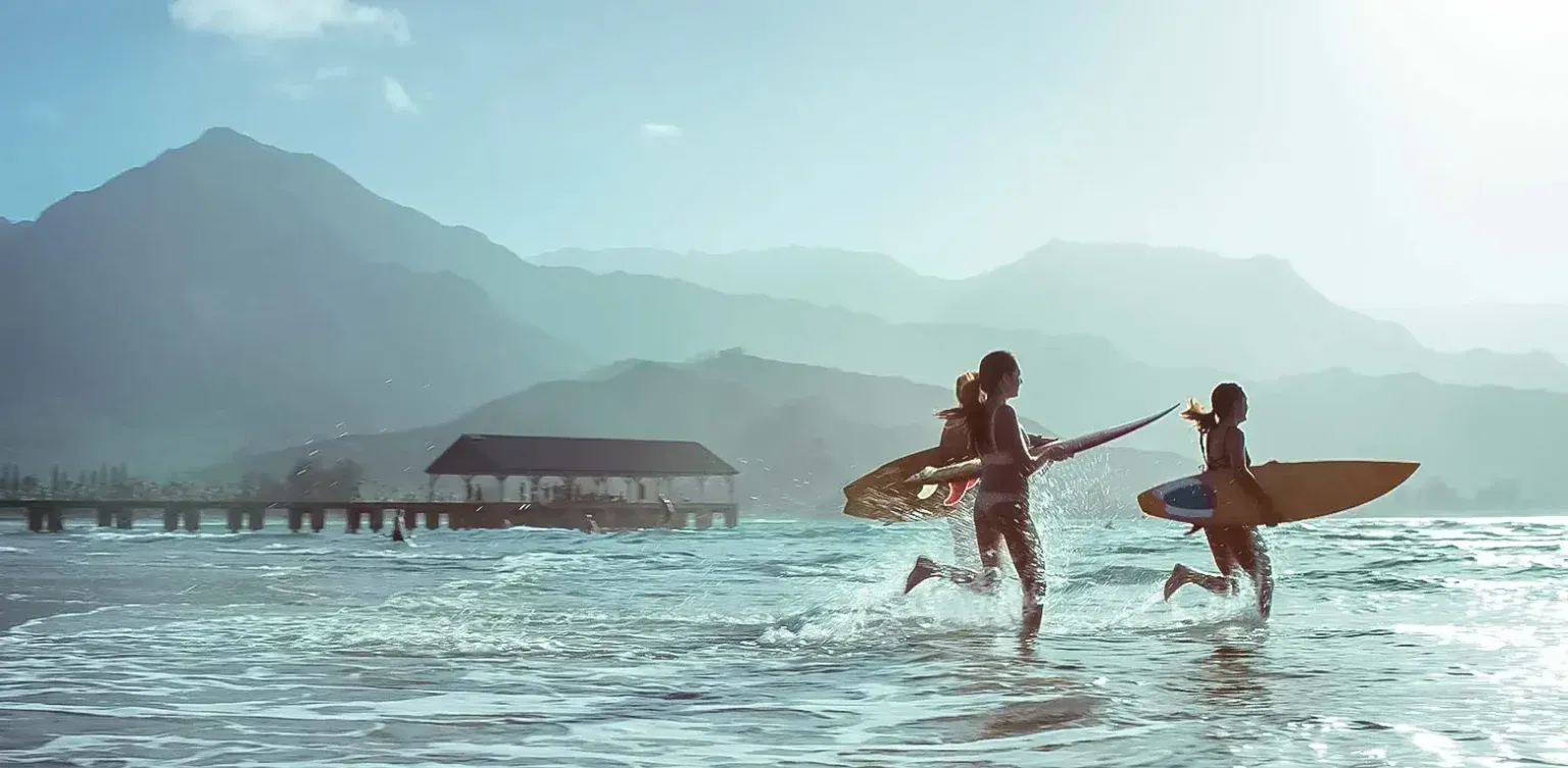 Two surfers run through shallow water at a misty beach with mountains in the background
