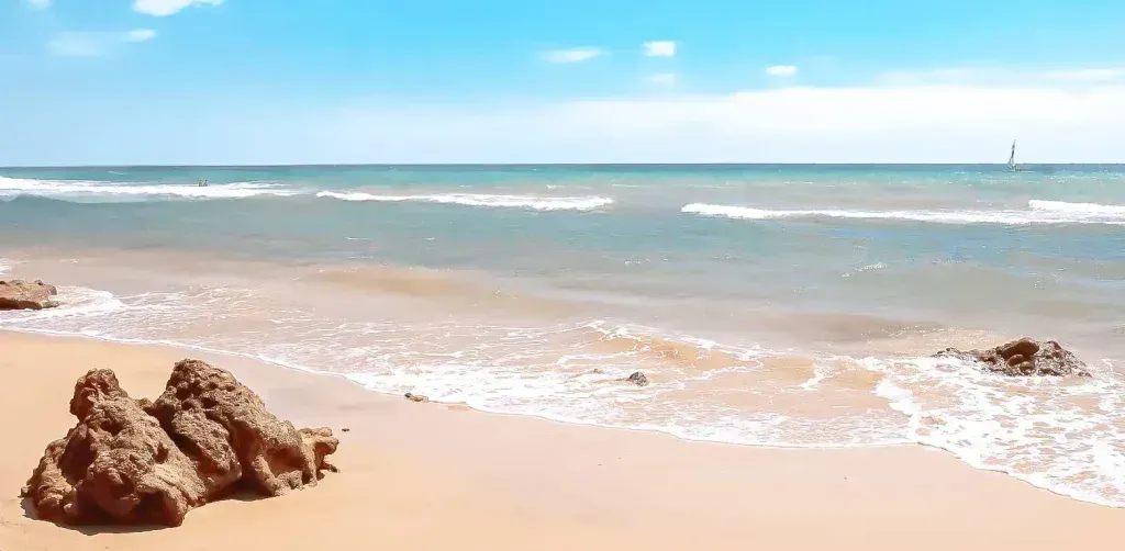 Sandy beach with gentle waves, rocks in the foreground, and a clear blue sky over calm water.