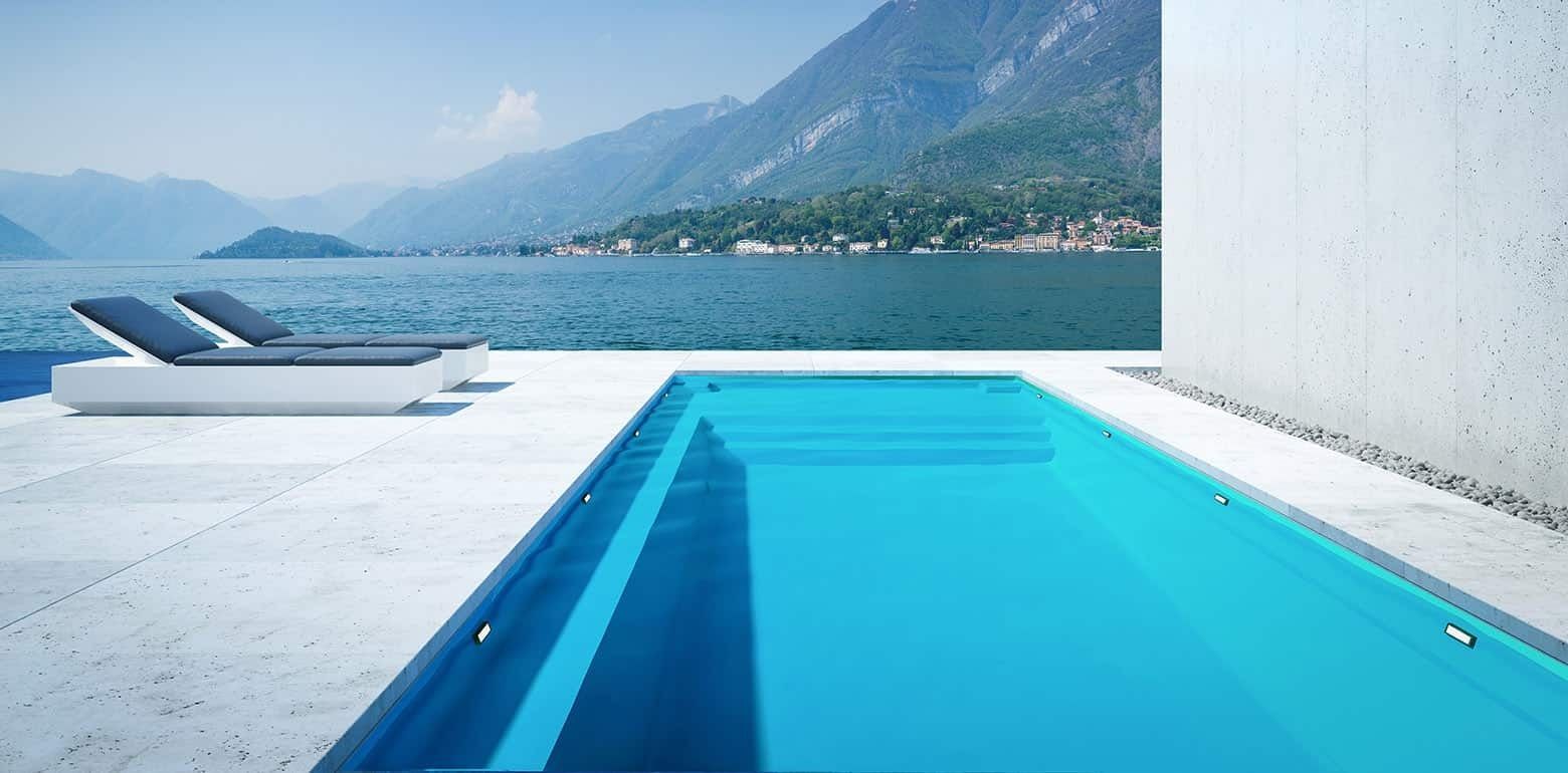 Modern infinity pool beside a lake and mountains, with white loungers and a bright blue water view