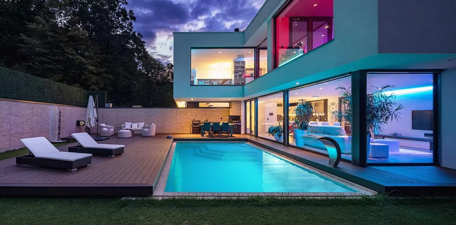 Modern luxury home with a lit patio, lounge chairs, and a glowing swimming pool at dusk