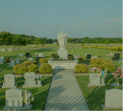 Cemetery with a central angel statue, flower arrangements, and headstones along a paved path under a clear sky