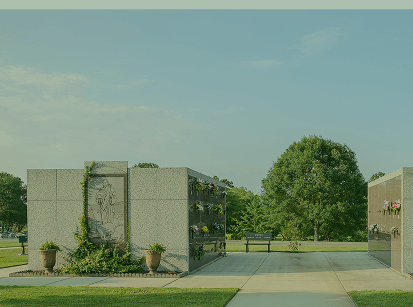 Stone entrance pillars beside a paved path, with green trees and a clear blue sky beyond