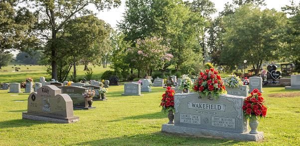 Gravestones in a green cemetery, with a gray headstone and red flower arrangements in the foreground