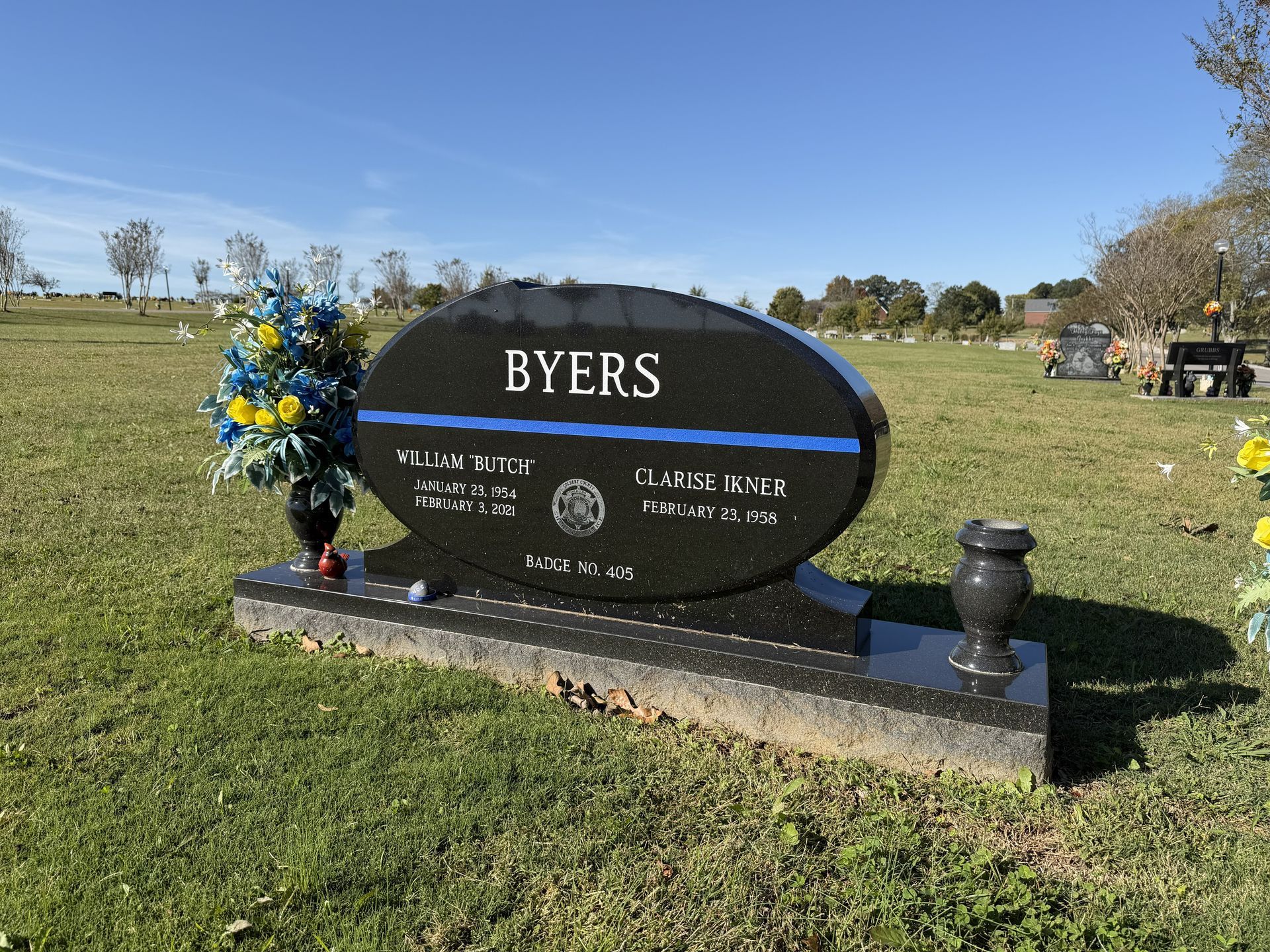Black oval memorial headstone engraved “BYERS” in a grassy cemetery, with blue and yellow flowers.