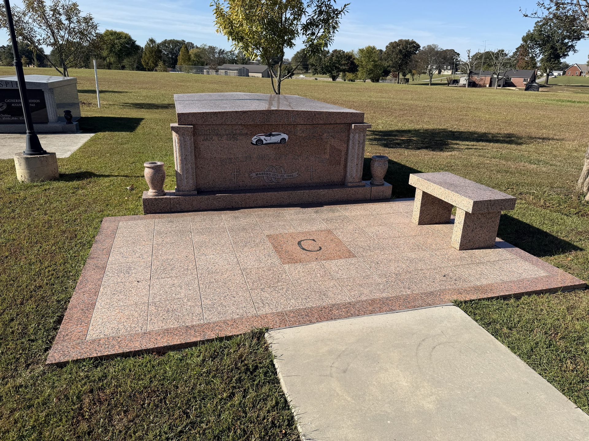 Gravestone with bench and flower vases in a grassy cemetery, with a concrete path in front.