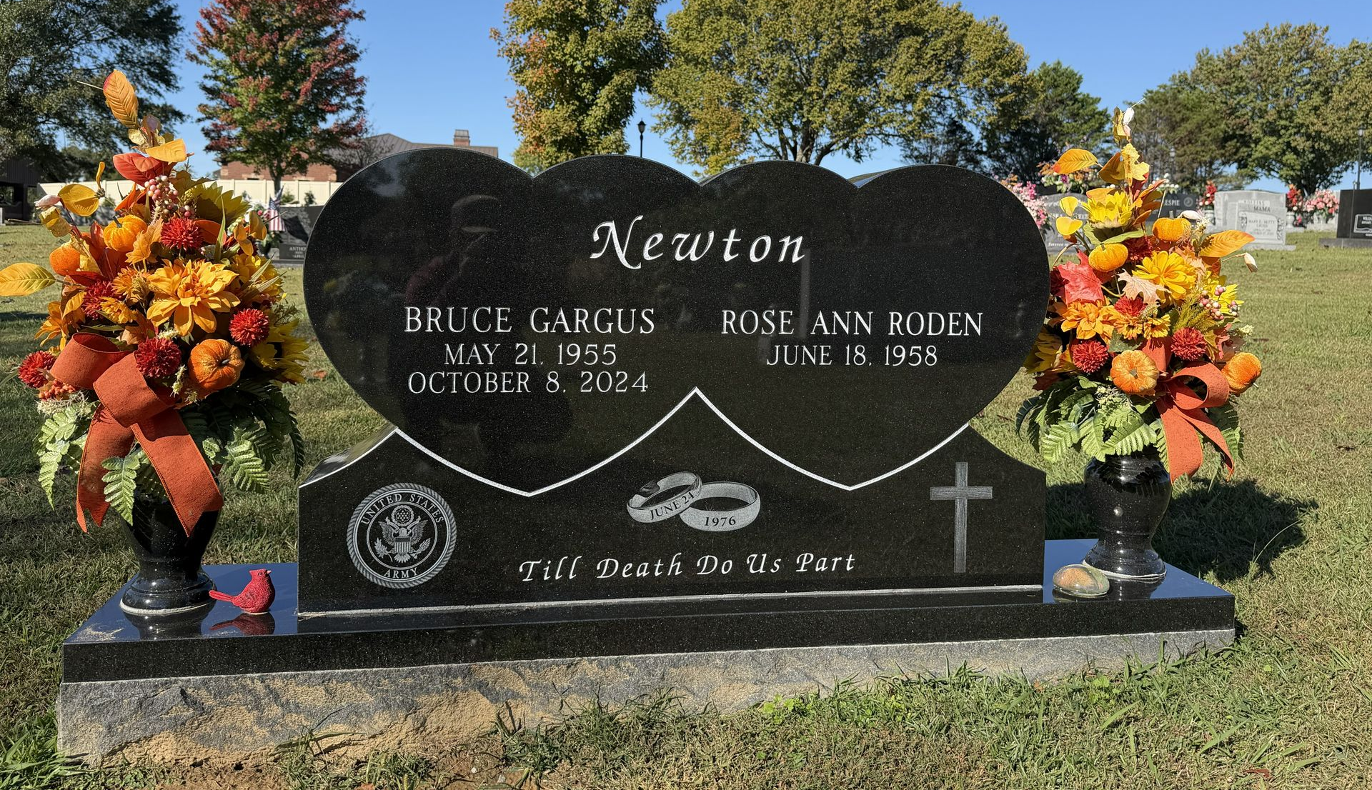 Black granite memorial headstone with heart-shaped engraving and flower arrangements on both sides