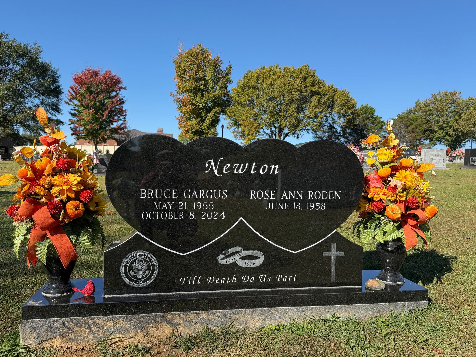 Black granite gravestone with flowers in a sunny cemetery, engraved “Newton” and two names.