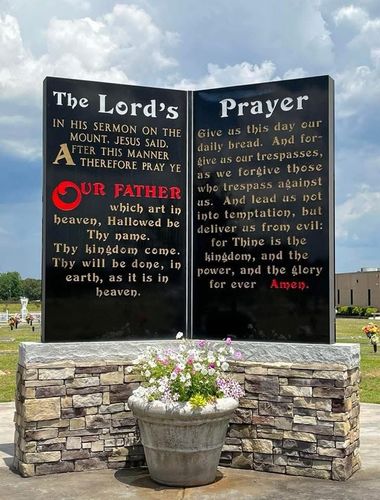 Stone religious sign with the Lord’s Prayer in a cemetery, set above a flower planter under a cloudy sky
