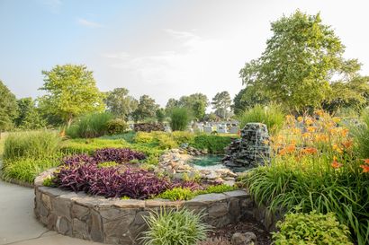 Stone-edged garden bed with colorful flowers, shrubs, and a small fountain in a landscaped park