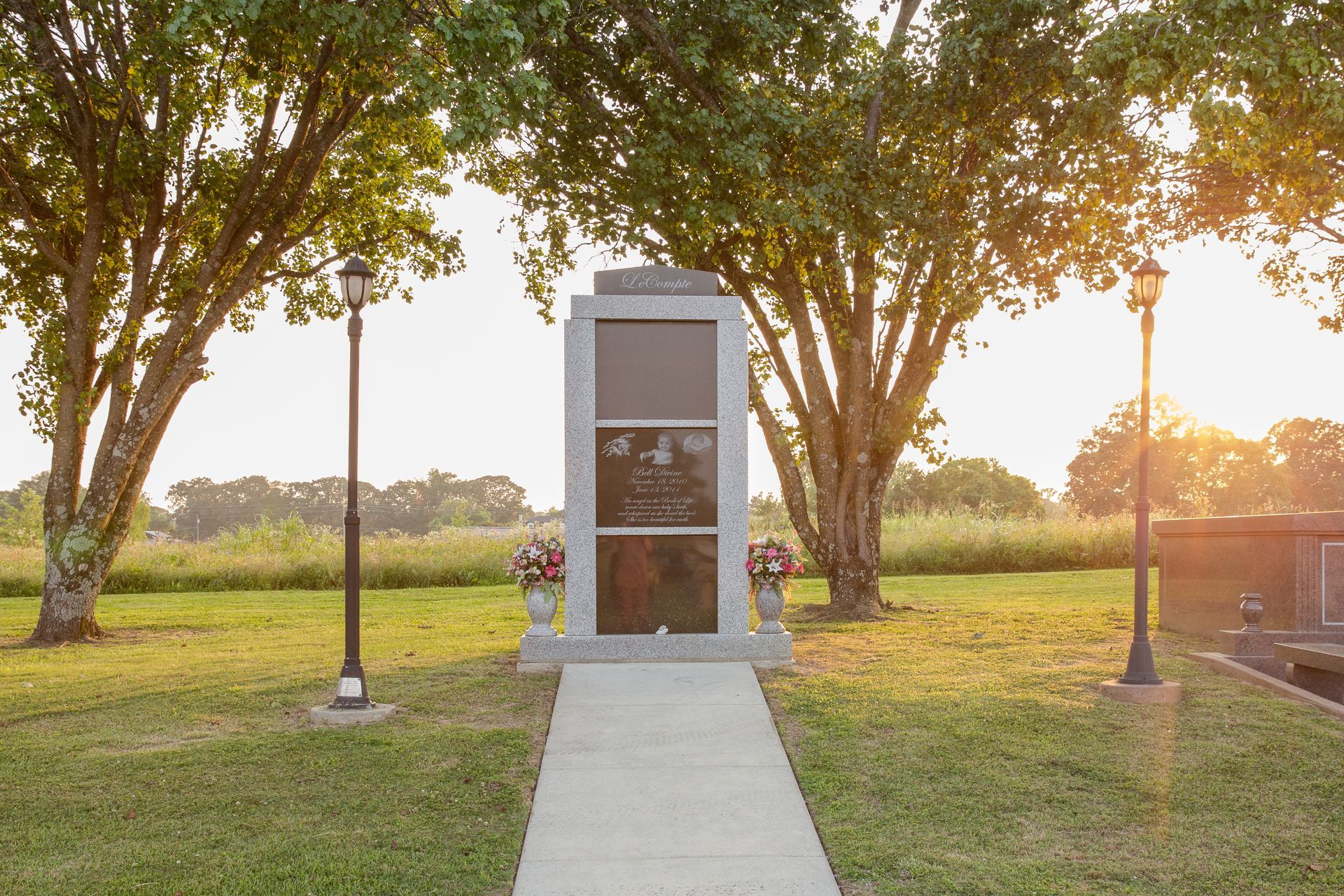 Memorial stone monument on a grassy path at sunset, framed by trees and lampposts.