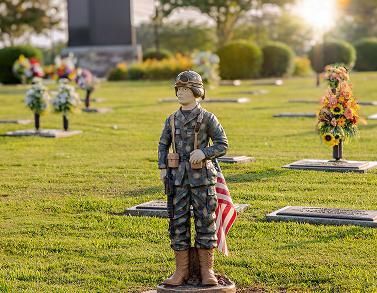 Soldier statue in camouflage holding a rifle, with an American flag in a sunlit cemetery