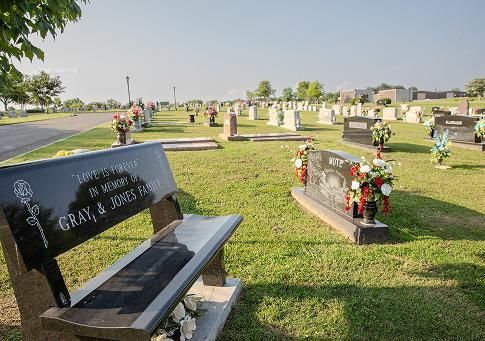 Cemetery with rows of headstones and flower arrangements under a clear sky, one black bench grave in foreground