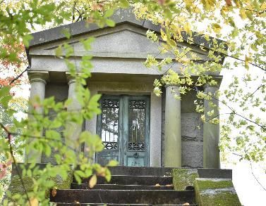 Stone mausoleum with columns and a blue door, partly obscured by green leaves