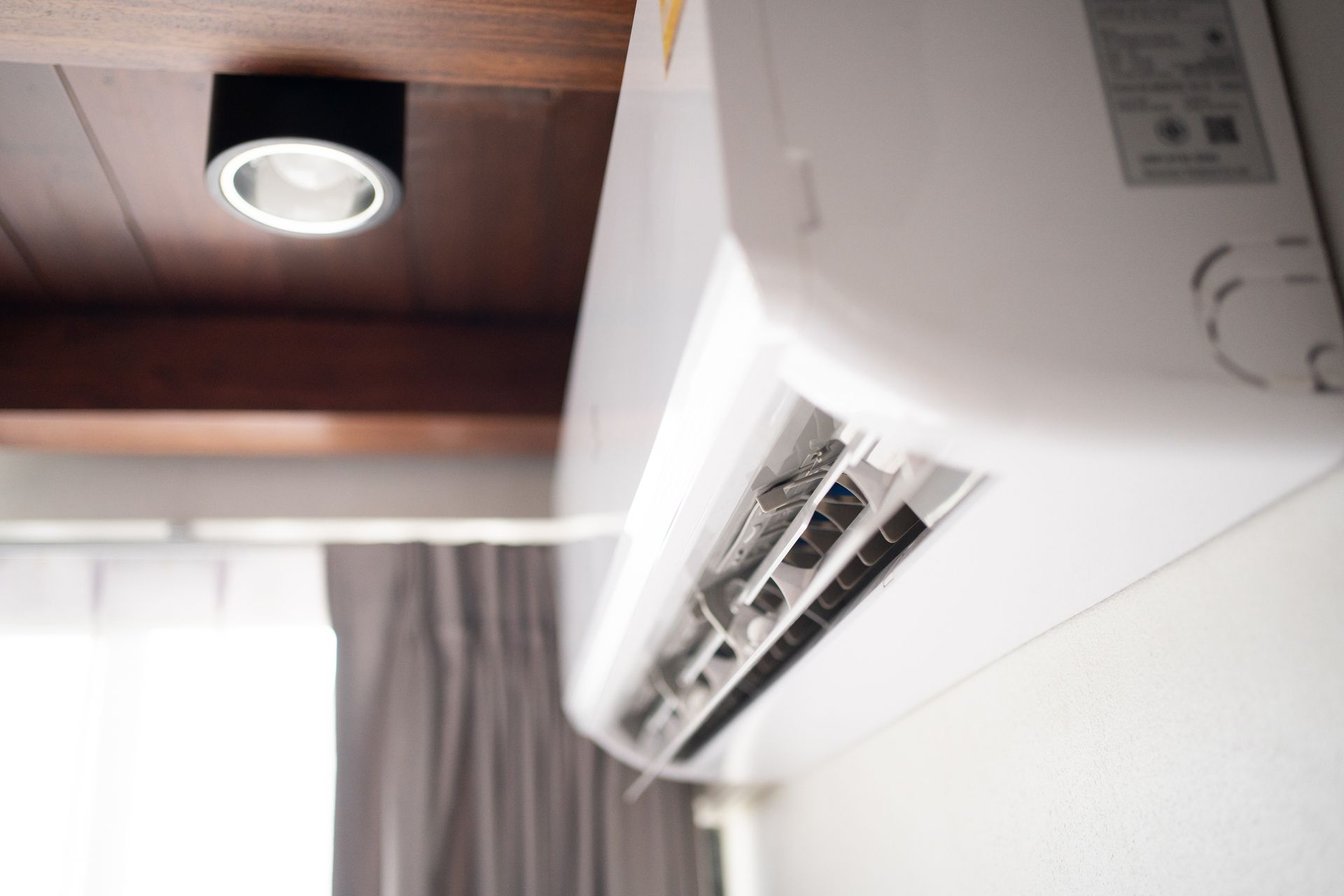 Condizionatore bianco montato su una parete in una stanza con soffitto in legno e tenda grigia.