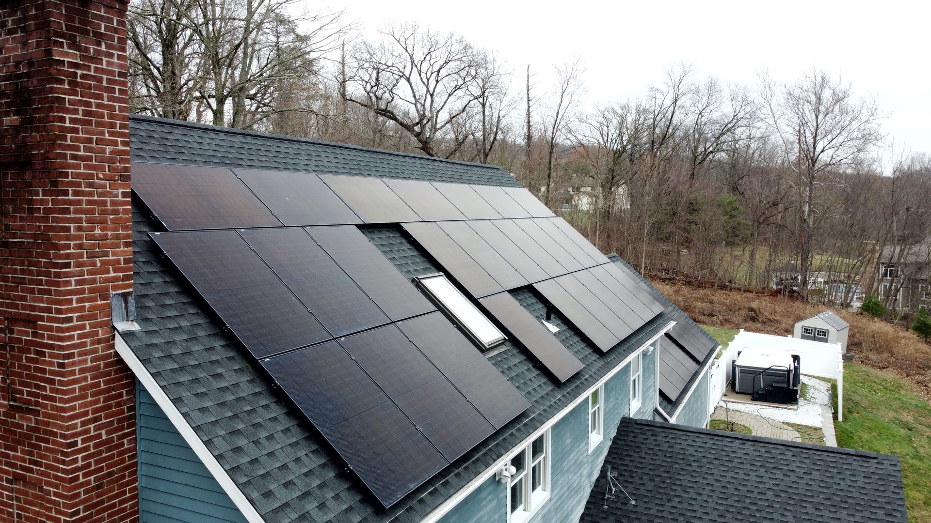 Solar panels installed on a dark shingled roof beside a wooded yard and white van