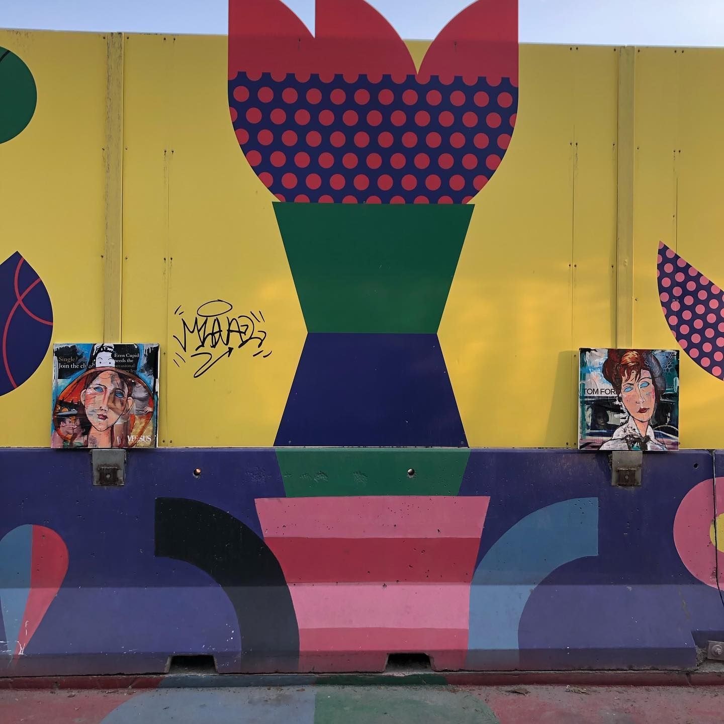 Due piccoli quadri incorniciati sono appesi a un murale giallo e viola caratterizzato da forme geometriche e motivi floreali stilizzati.