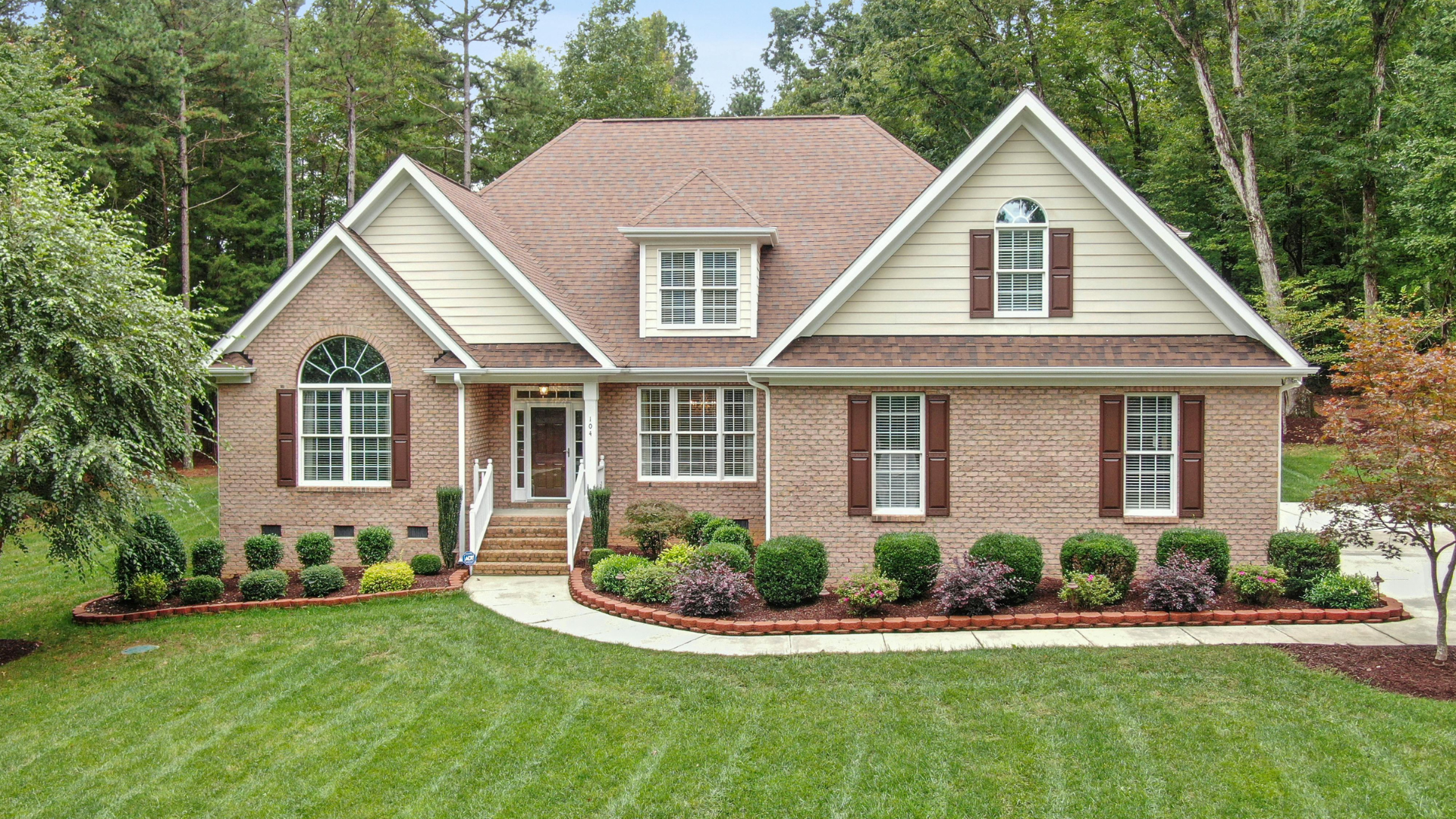 Two-story brick and siding house with a front lawn, sidewalk, and lush green trees in the background.