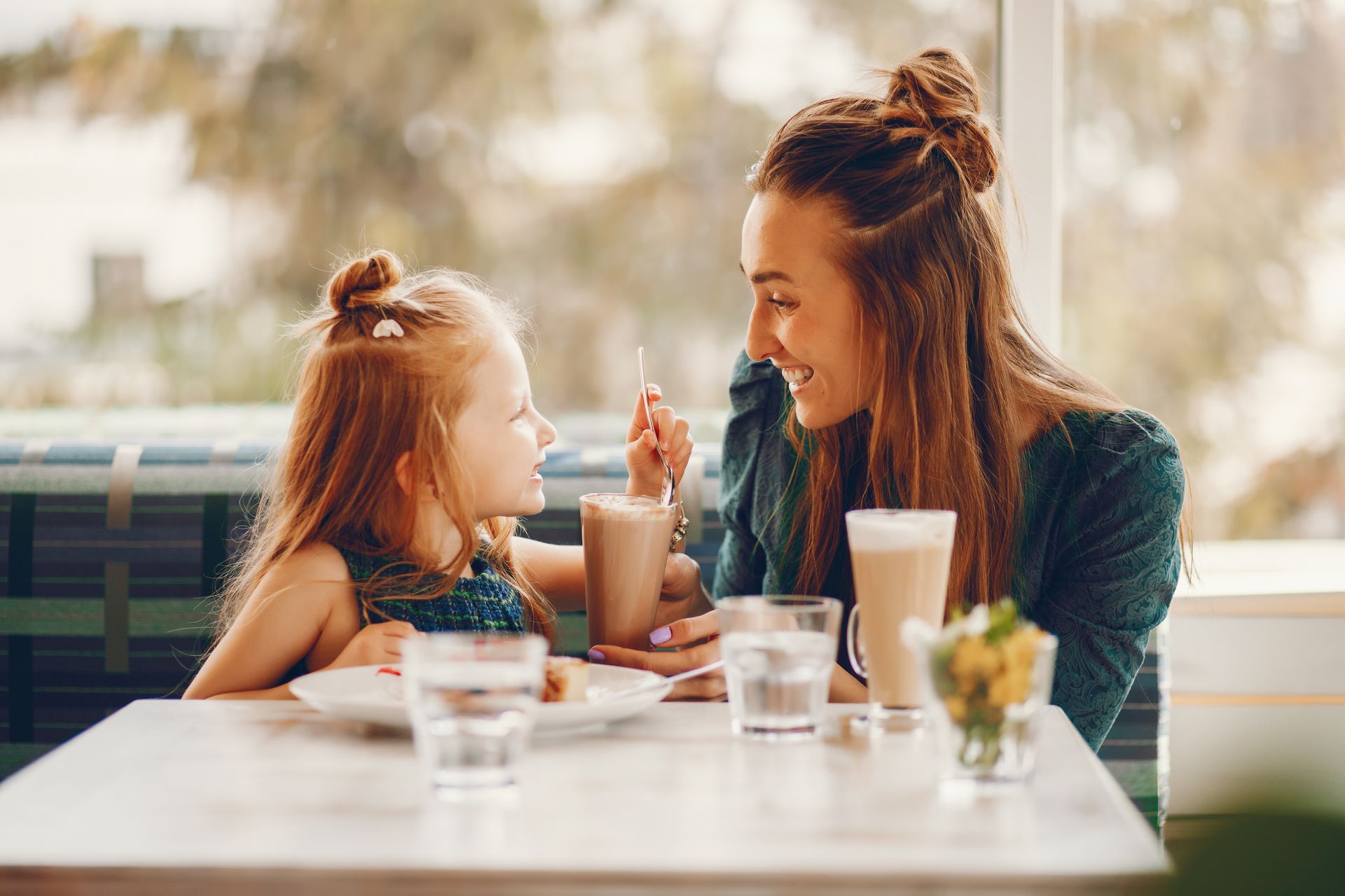 Woman smiles at child, both at a table in a cafe. They share a drink.