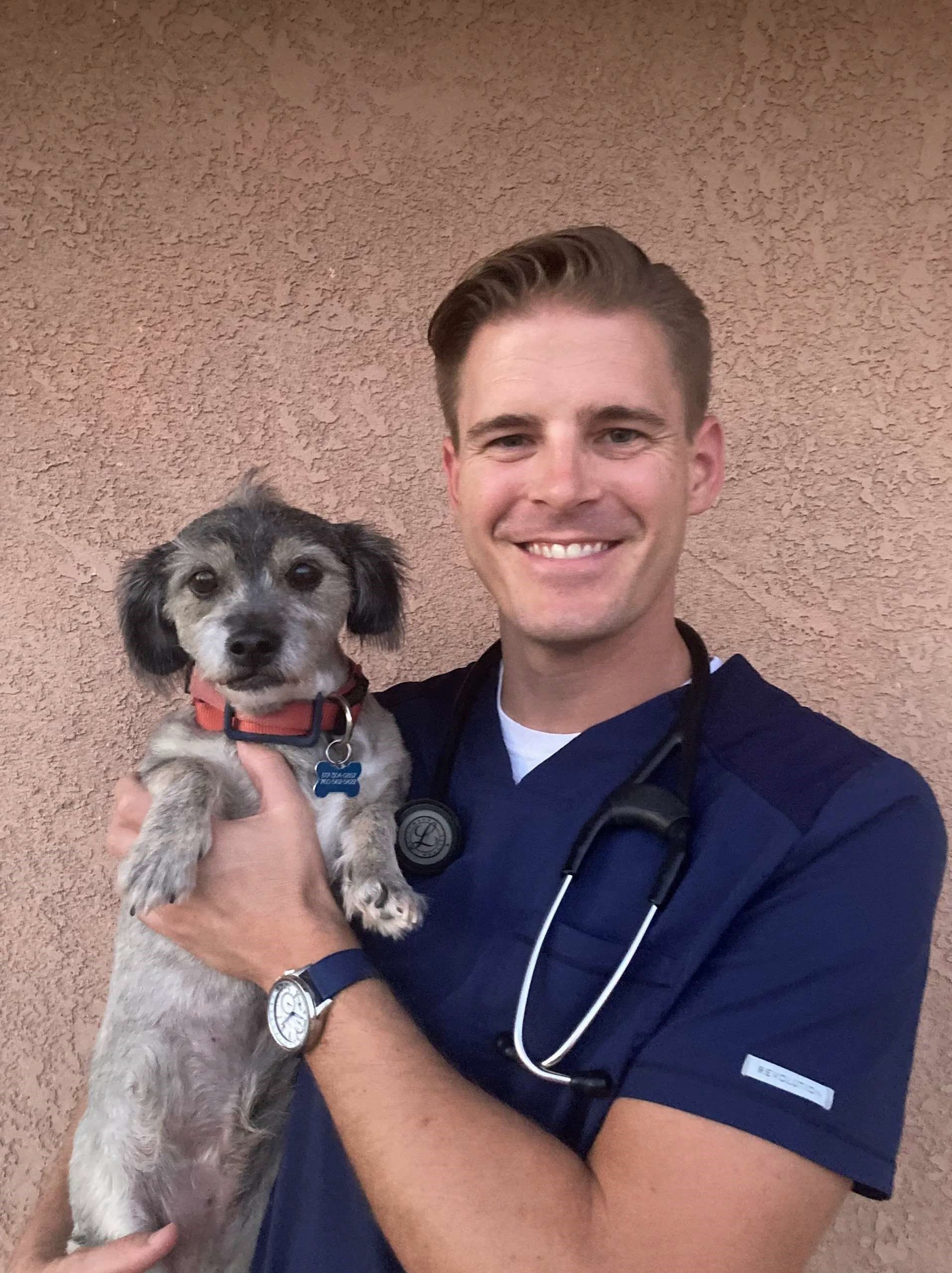 Veterinarian in blue scrubs holds a small dog; both are smiling. Background is a textured, light brown wall. Veterinarian in blue scrubs holds a small dog; both are smiling. Background is a textured, light brown wall.