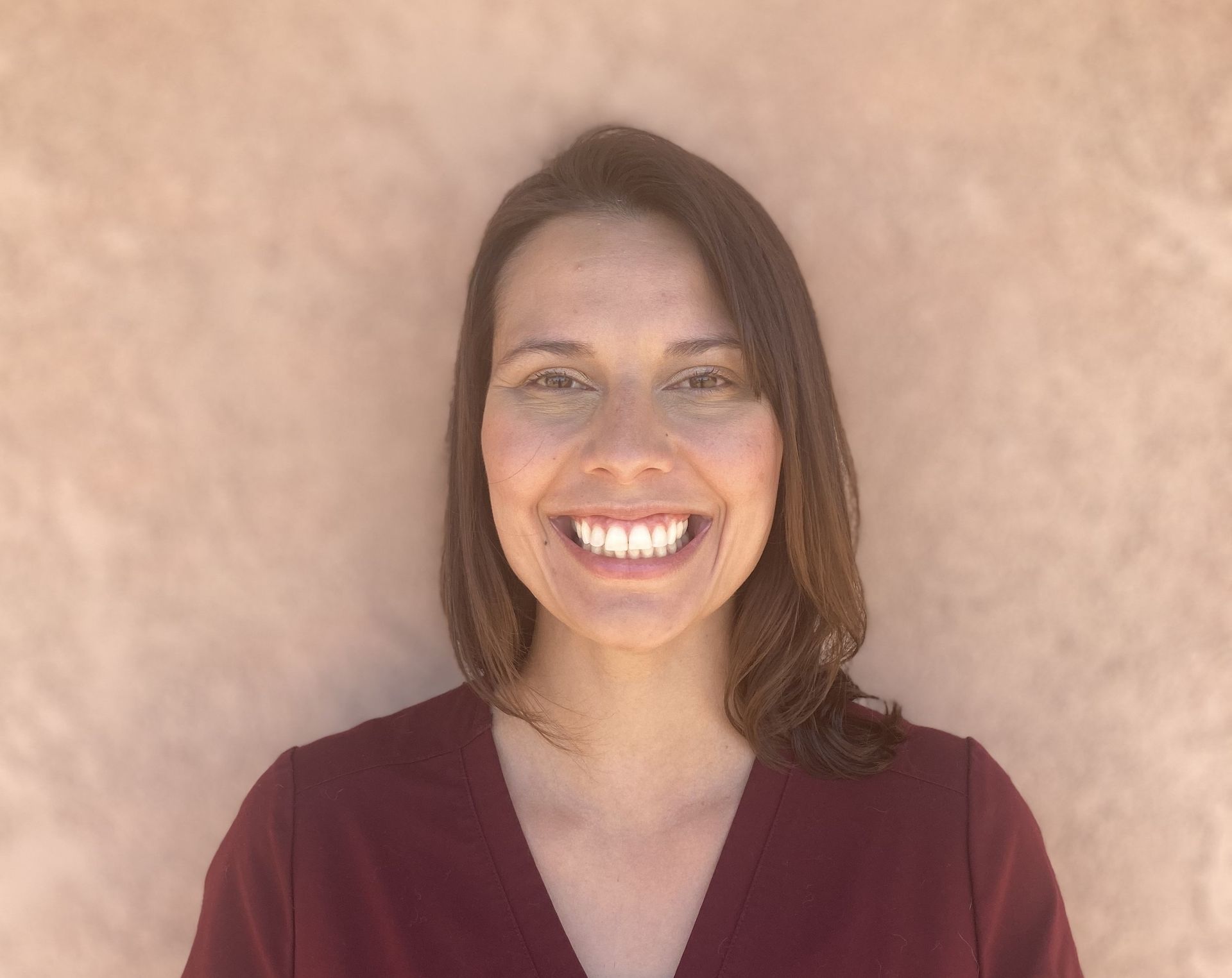 Woman with shoulder-length brown hair smiles at the camera, wearing a dark burgundy top, against a peach-colored wall. Woman with shoulder-length brown hair smiles at the camera, wearing a dark burgundy top, against a peach-colored wall.