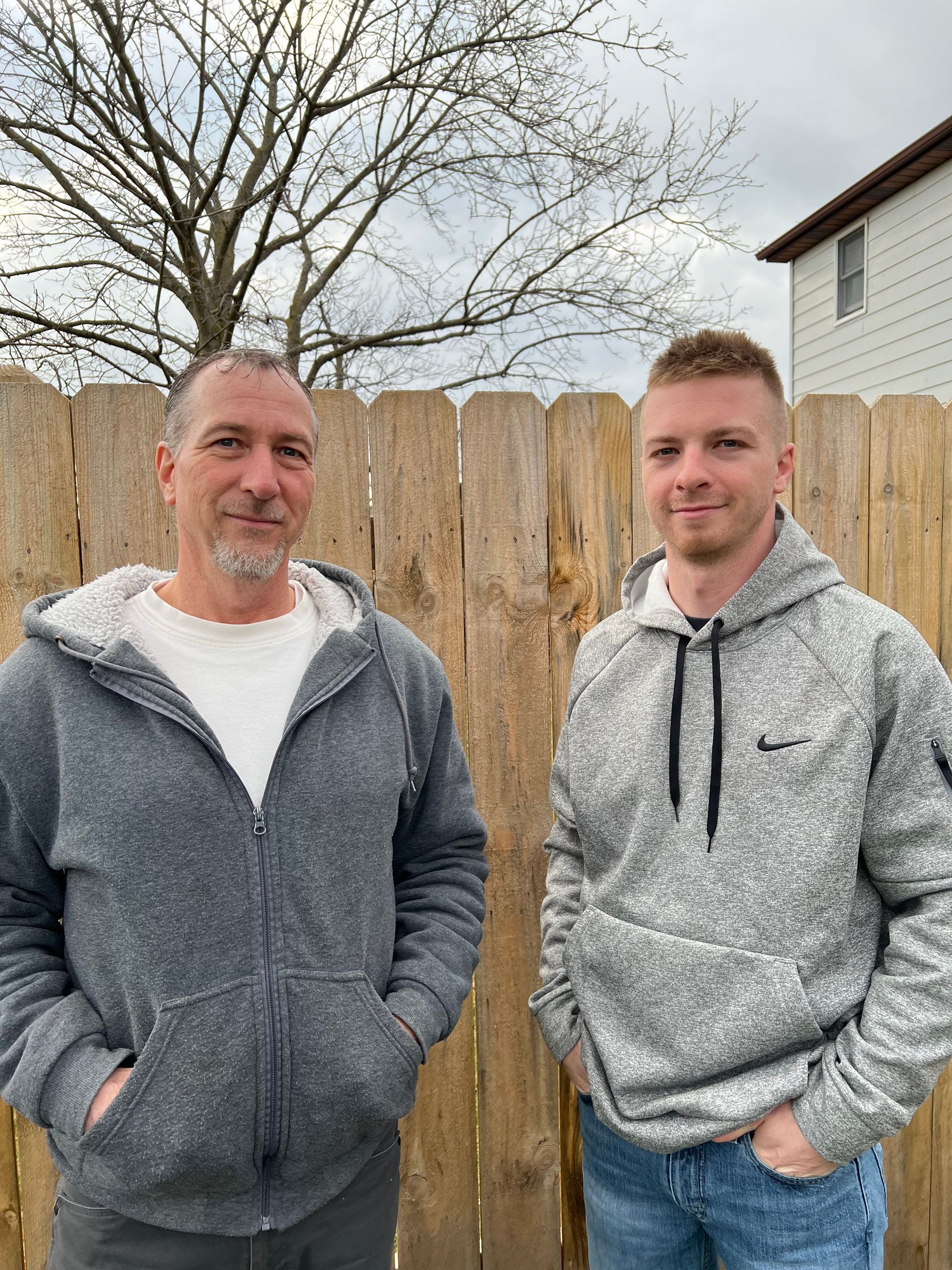 Two men, Jeff & Shane standing side by side against a wooden fence, smiling for the camera in casual attire