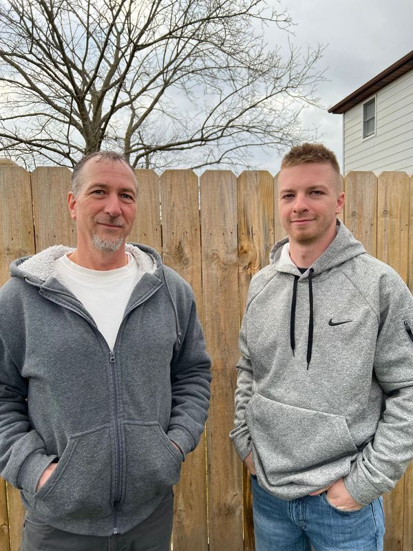 two men are standing next to each other in front of a wooden fence .